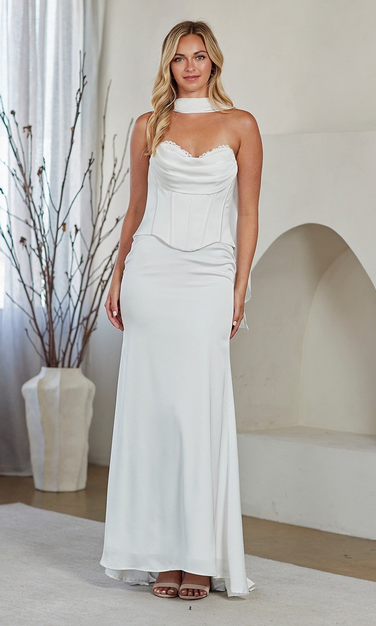A woman stands indoors wearing the Amelia Couture 6137, a long white prom dress with a strapless corset bodice, choker-style neckline, and an elegantly flowing floor-length skirt.